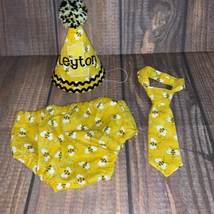 May include: A yellow and black bee-themed baby outfit set with a party hat, diaper cover, and tie. The hat has a black pom-pom on top and says "Leyton" on the front.