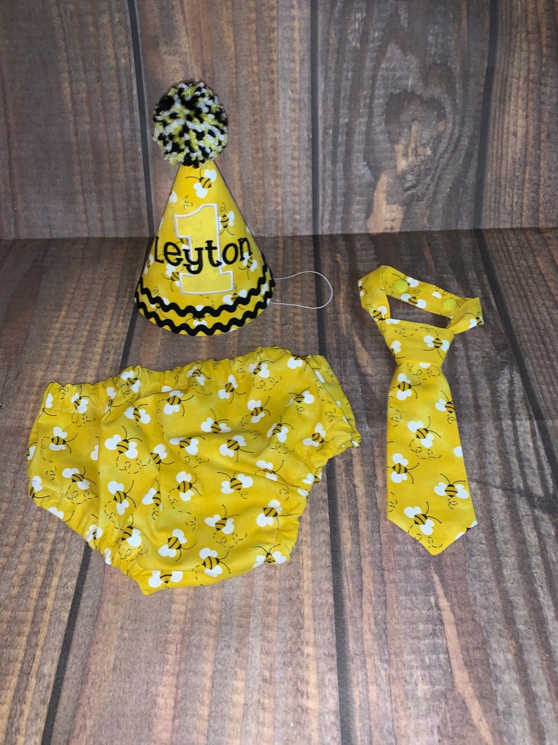 May include: A yellow and black bee-themed baby outfit set with a party hat, diaper cover, and tie. The hat has a black pom-pom on top and says "Leyton" on the front.