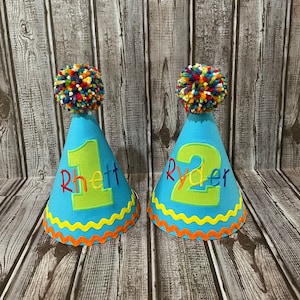 May include: Two blue party hats with colorful pom-poms on top. The hats have yellow and orange trim and say "Rhett 1" and "Ryder 2" in green letters.