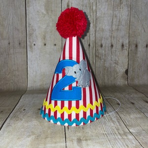 May include: Red and white striped party hat with a blue number 2 and a grey elephant applique. The hat has a red pom pom on top and a yellow and blue trim.