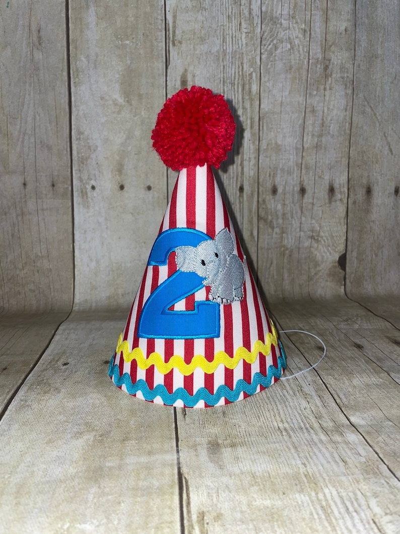 May include: Red and white striped party hat with a blue number 2 and a grey elephant applique. The hat has a red pom pom on top and a yellow and blue trim.