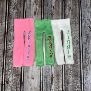 May include: Three pairs of elf legs in pink, green, and white with embroidered names "Sprinkles", "Buddy", and "Jingle" on each pair.
