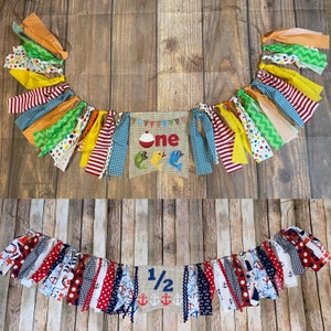Op de afbeelding: Een kleurrijk stoffen banner met het woord "one" op een jute achtergrond. De banner is versierd met vissen, een dobber en rode, gele, blauwe en groene stofstroken. Een tweede banner met de tekst "1/2" op een jute achtergrond is versierd met ankers en rode, witte en blauwe stofstroken.