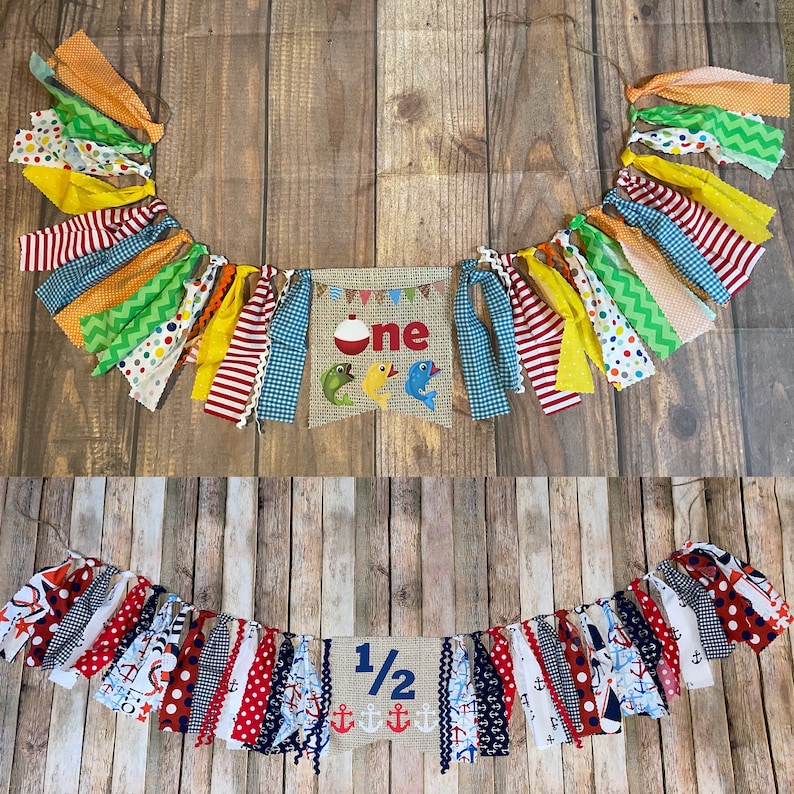 Op de afbeelding: Een kleurrijk stoffen banner met het woord "one" op een jute achtergrond. De banner is versierd met vissen, een dobber en rode, gele, blauwe en groene stofstroken. Een tweede banner met de tekst "1/2" op een jute achtergrond is versierd met ankers en rode, witte en blauwe stofstroken.