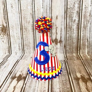 May include: A red and white striped party hat with a blue number 3 and the name Luca embroidered on the front. The hat has a yellow and blue trim and a red, yellow, and blue pom-pom on top.