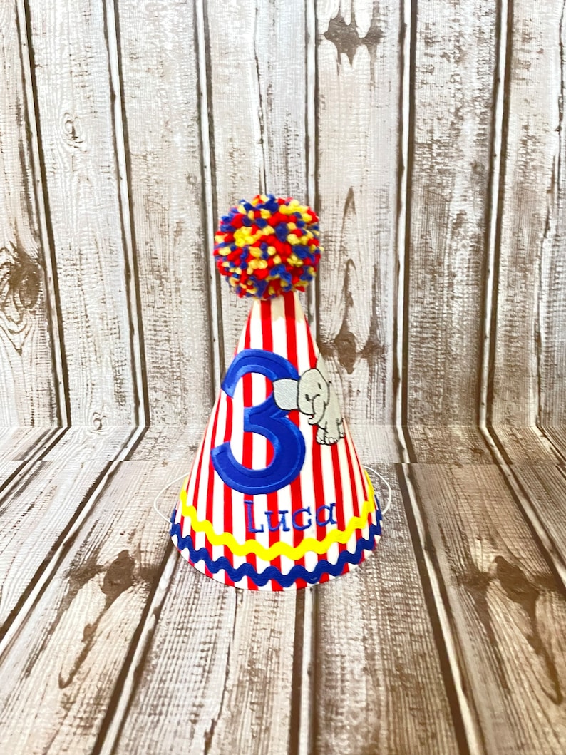 May include: A red and white striped party hat with a blue number 3 and the name Luca embroidered on the front. The hat has a yellow and blue trim and a red, yellow, and blue pom-pom on top.
