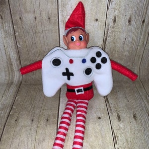 May include: A Christmas elf dressed as a video game controller. The elf is wearing a white and black controller costume with red and white striped pants and a red hat.