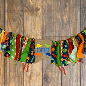 Op de afbeelding: Een kleurrijk stoffen banner met dinosaurusontwerpen en het woord "one" in blauw op een jute achtergrond. De banner is aan een wit koord geregen en heeft een verscheidenheid aan kleuren, waaronder rood, groen, geel, oranje en blauw.