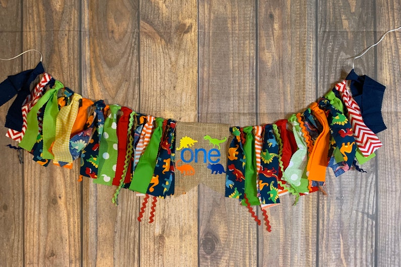 Op de afbeelding: Een kleurrijk stoffen banner met dinosaurusontwerpen en het woord "one" in blauw op een jute achtergrond. De banner is aan een wit koord geregen en heeft een verscheidenheid aan kleuren, waaronder rood, groen, geel, oranje en blauw.