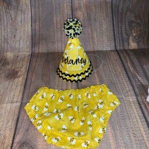 May include: Yellow and black bee-themed baby outfit with a matching party hat. The hat has a black pom-pom and the name "Delaney" embroidered on the front.