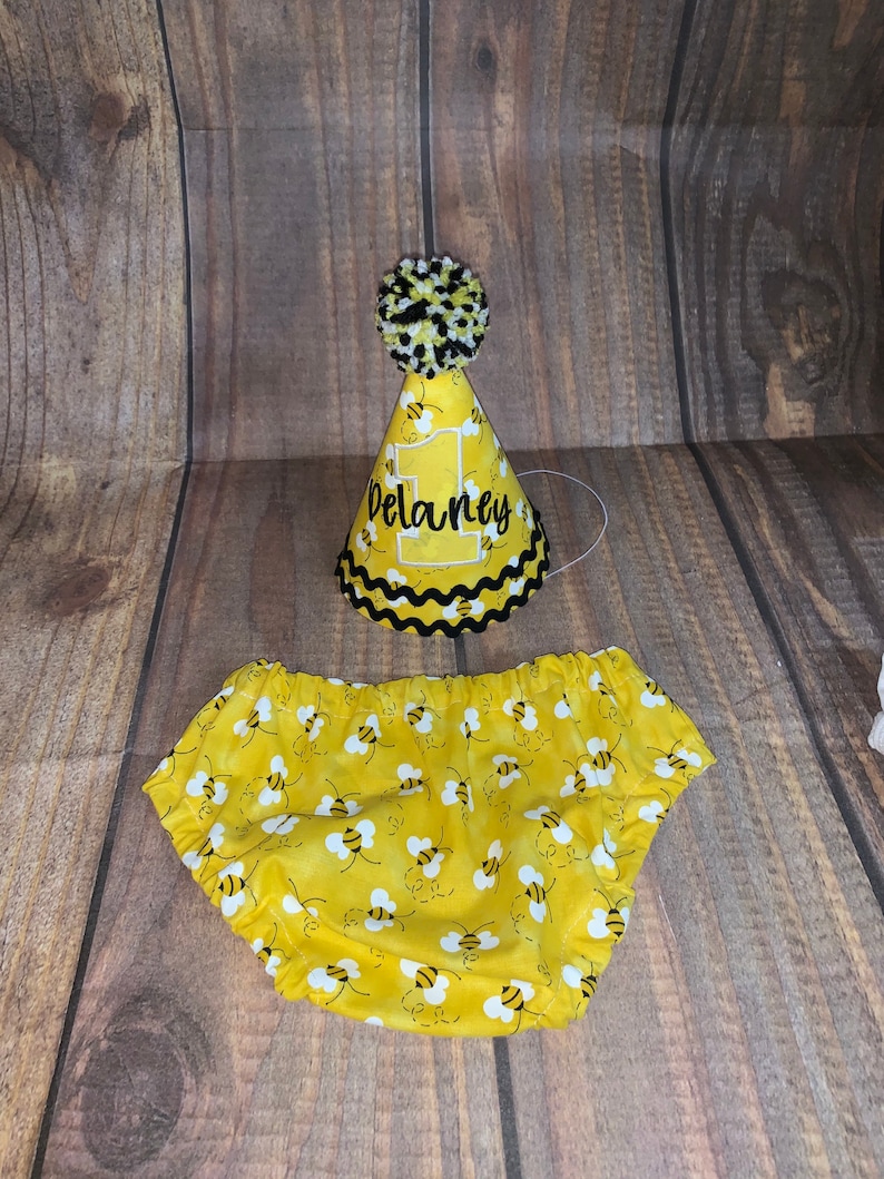 May include: Yellow and black bee-themed baby outfit with a matching party hat. The hat has a black pom-pom and the name "Delaney" embroidered on the front.