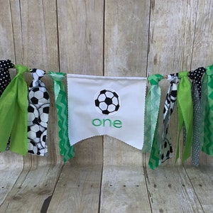 Op de afbeelding: Een groene, zwarte en witte stoffen banner met een witte vlag in het midden waarop "one" staat en een groene voetbal is afgebeeld. De banner is versierd met groene, zwarte en witte stoffen kwastjes.