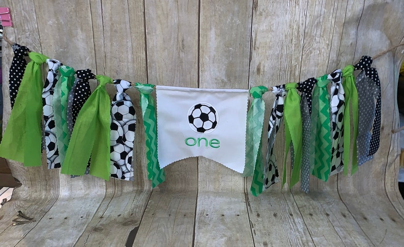 Op de afbeelding: Een groene, zwarte en witte stoffen banner met een witte vlag in het midden waarop "one" staat en een groene voetbal is afgebeeld. De banner is versierd met groene, zwarte en witte stoffen kwastjes.