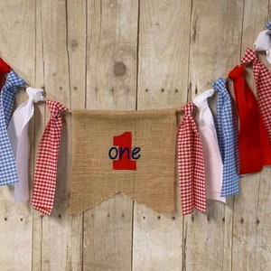 Op de afbeelding: Een banner van jute met het woord "one" in rood en blauw borduurwerk. De banner is bespannen met rode, witte en blauwe gingham- en effen stof lint streamers.