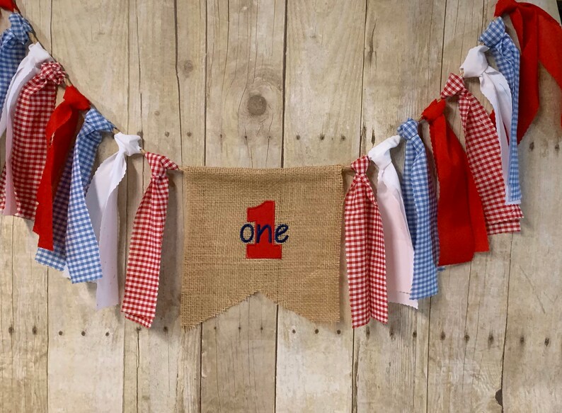 Op de afbeelding: Een banner van jute met het woord "one" in rood en blauw borduurwerk. De banner is bespannen met rode, witte en blauwe gingham- en effen stof lint streamers.