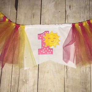 Op de afbeelding: Een witte banner met een roze nummer &eacute;&eacute;n en een gele zon met een glimlach. De banner is versierd met rood en geel tule.
