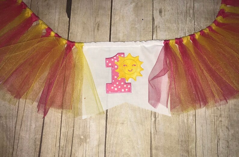Op de afbeelding: Een witte banner met een roze nummer &eacute;&eacute;n en een gele zon met een glimlach. De banner is versierd met rood en geel tule.