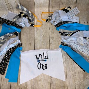 Op de afbeelding: Een witte stoffen banner met de tekst "Wild One" en een pijl die naar rechts wijst. De banner is versierd met blauwe, grijze en zwarte stoffen streamers.