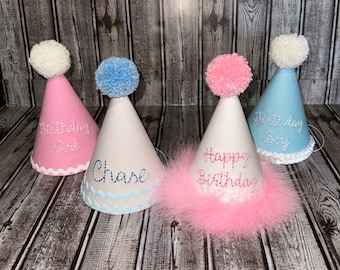 May include: Four party hats with pom-poms and different colored trims. The hats are white with pink and blue embroidery. One hat says "Happy Birthday", one says "First Birthday Girl", one says "First Birthday Boy", and one says "Chase".
