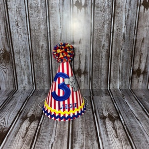 May include: Red and white striped party hat with a blue number 3, a small grey elephant, and the name Luca in blue lettering. The hat has a yellow and red pom-pom on top.