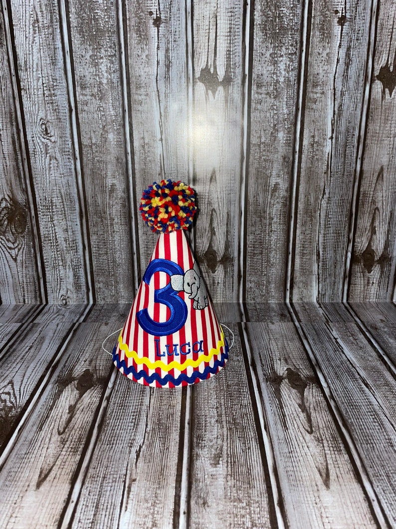 May include: Red and white striped party hat with a blue number 3, a small grey elephant, and the name Luca in blue lettering. The hat has a yellow and red pom-pom on top.