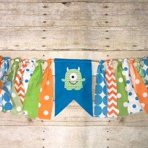 Op de afbeelding: Een blauwe en groene monsterbanner met een groen monstergezicht in het midden. De banner is gemaakt van stofresten met verschillende patronen, waaronder stippen, chevron en effen kleuren. De banner is aan een touwtje geregen.