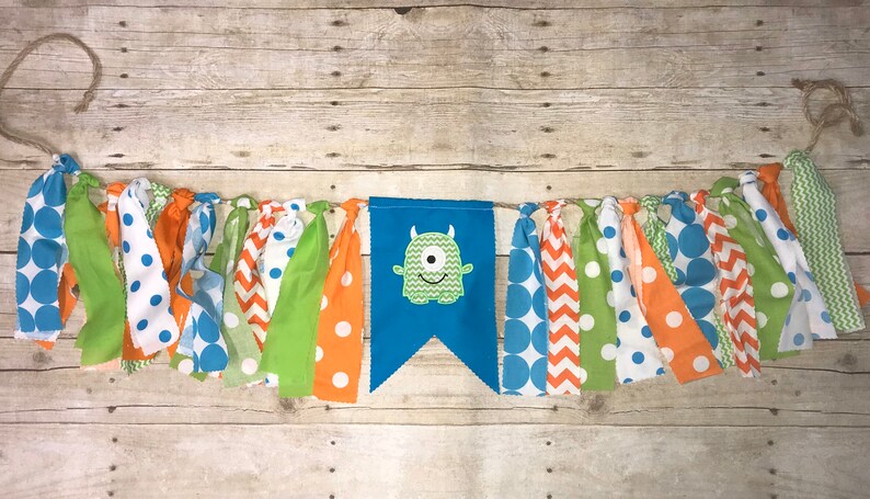Op de afbeelding: Een blauwe en groene monsterbanner met een groen monstergezicht in het midden. De banner is gemaakt van stofresten met verschillende patronen, waaronder stippen, chevron en effen kleuren. De banner is aan een touwtje geregen.