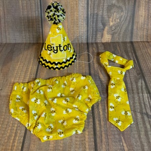 May include: A yellow and black bee-themed baby outfit set with a party hat, diaper cover, and tie. The hat has a black pom-pom on top and says "Leyton" on the front.