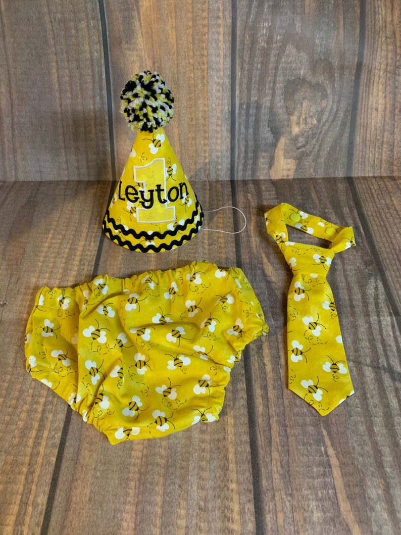 May include: A yellow and black bee-themed baby outfit set with a party hat, diaper cover, and tie. The hat has a black pom-pom on top and says "Leyton" on the front.