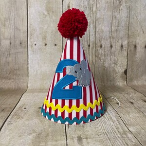May include: Red and white striped party hat with a red pom-pom on top. The hat has a blue number "2" and a grey elephant applique. The hat has a yellow and teal trim.