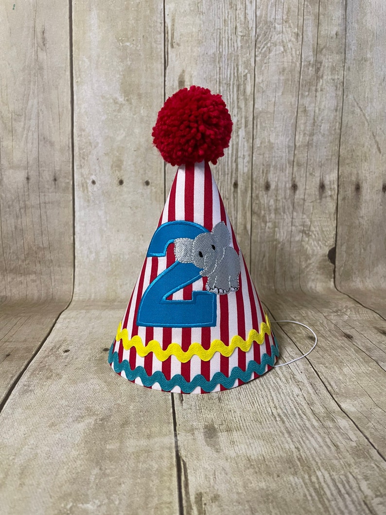 May include: Red and white striped party hat with a red pom-pom on top. The hat has a blue number "2" and a grey elephant applique. The hat has a yellow and teal trim.