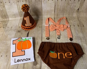 Può includere: Un completo per il primo compleanno di un bambino a tema zucca, di colore marrone e arancione. Il completo comprende una cravatta a farfalla in gingham marrone e arancione, bretelle, un pannolino marrone con la parola "uno" e una zucca, e una tutina bianca con una grande zucca arancione e il numero "uno" con il nome "Lennox" sotto. Il completo include anche un cappello da festa in gingham marrone e arancione con un pon pon marrone in cima.