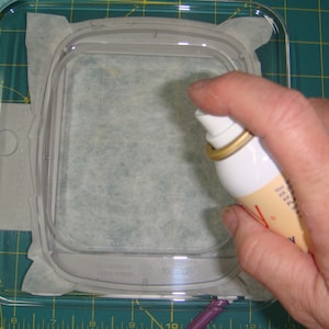 May include: A hand holding a spray bottle over a white fabric in a clear plastic embroidery hoop. The fabric is in a clear plastic container on a green cutting mat.
