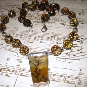 May include: A brown and gold gemstone necklace with a rectangular pendant featuring a dendritic agate stone. The necklace is made with faceted beads and silver accents.