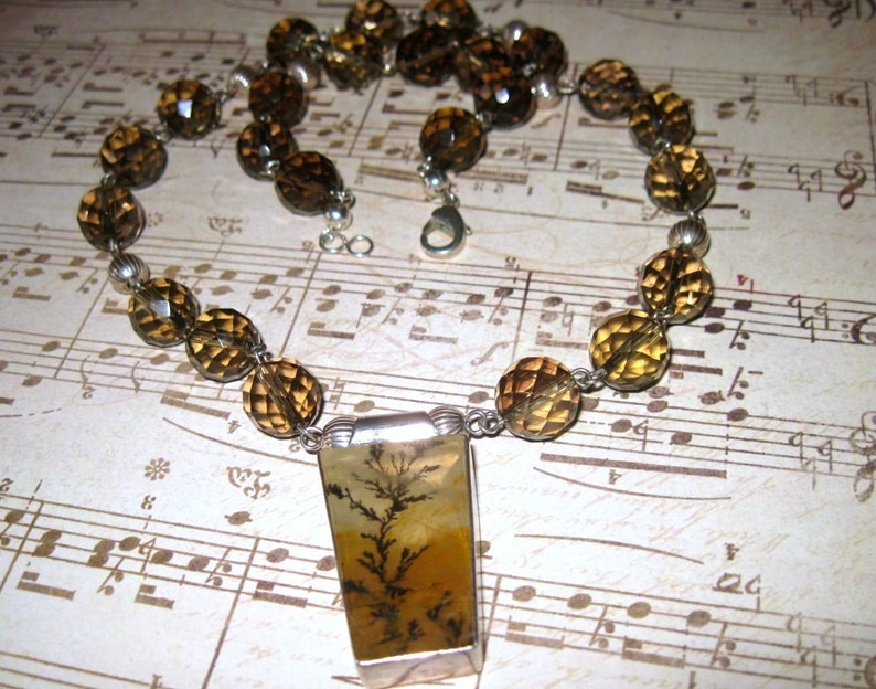 May include: A brown and gold gemstone necklace with a rectangular pendant featuring a dendritic agate stone. The necklace is made with faceted beads and silver accents.