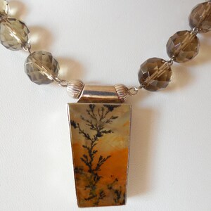 May include: A silver necklace with a rectangular pendant featuring a dendritic agate stone with brown, black, and orange patterns. The necklace is made with faceted, smoky quartz beads.