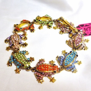 May include: A gold-tone bracelet with a chain link design featuring seven colorful frog charms. Each frog is adorned with rhinestones in various colors, including green, blue, pink, purple, and orange.