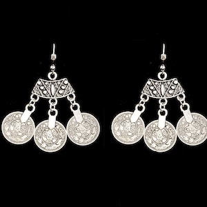 May include: Silver-toned dangle earrings featuring a decorative top with three coin-shaped pendants. The coins have intricate embossed designs. The earrings are displayed against a black background.