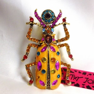 May include: A jeweled beetle brooch with a golden body, adorned with colorful rhinestones in shades of orange, purple, blue, and red. The brooch has intricate details and a decorative design.