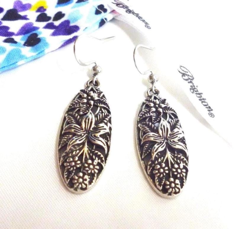 Pewter tone earrings with a raised floral design with a Brighton dustbag. They have a flower on in the center that looks like a lily. They have hook attachments.