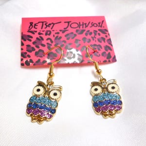 May include: A pair of gold-tone hook earrings featuring owl-shaped pendants with cream-colored faces and glittery blue, purple, and pink bodies. The earrings are attached to a pink display card with a black leopard print and the text Betsey Johnson.