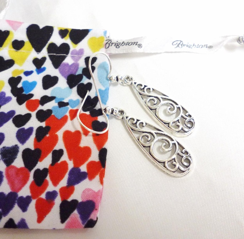 A pair of teardrop shaped, silver plated earrings. They are designed with openwork filigree in a swirling pattern. They are displayed with a multi colored heart themed Brighton cloth bag.