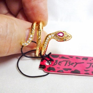 May include: Gold-tone snake ring with a red gemstone eye and clear crystal accents. The ring is wrapped around a finger and the brand name "Betsey Johnson" is visible on a pink tag.