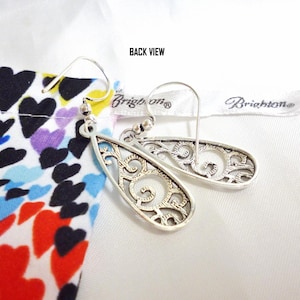 A back view of the earrings, showing the pattern in reverse, with a texured finish on the back. They have French hook attachments. The photo is labeled, 'Back View'. They are designed with openwork filigree in a swirling pattern. They are displayed with a multi colored heart themed Bright bag.