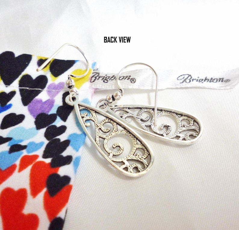 A back view of the earrings, showing the pattern in reverse, with a texured finish on the back. They have French hook attachments. The photo is labeled, 'Back View'. They are designed with openwork filigree in a swirling pattern. They are displayed with a multi colored heart themed Bright bag.