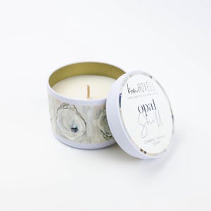 May include: A white tin candle with a white lid. The candle has a design of oyster shells on the side. The lid has the text "Kim Hovelle Hand Crafted Soy Wax Candles Opal Shell Cypress Orange Cedar".