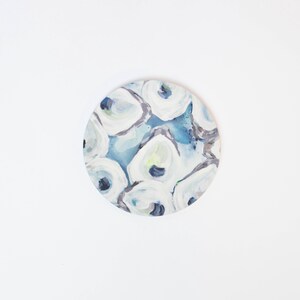 May include: Round, abstract art piece with a coastal theme. The artwork features a pattern of white, blue, and gray brushstrokes, resembling oyster shells. The background is a mix of light and dark blues.