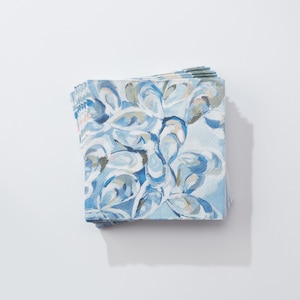 May include: A stack of square paper napkins with a blue and white abstract design. The pattern features a watercolor-style depiction of oyster shells in shades of blue, gray, and beige. The napkins are stacked neatly on a white surface.