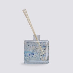 May include: A clear glass bottle diffuser with a blue and white watercolor design of seashells. The bottle has a label that reads "Kim Novelli Underwater Water & Air Collection Diffuser". The diffuser has wooden sticks in it.