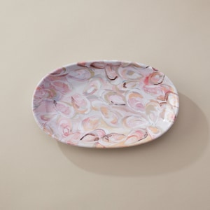 May include: An oval serving platter with a white background and a pink and beige abstract pattern of oyster shells.